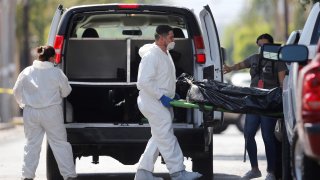 Tres peritos forenses, dos con trajes blancos, trasladan el cuerpo de una persona en una especie de camilla que bajaron de una camioneta negra.