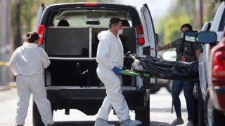 Tres peritos forenses, dos con trajes blancos, trasladan el cuerpo de una persona en una especie de camilla que bajaron de una camioneta negra.