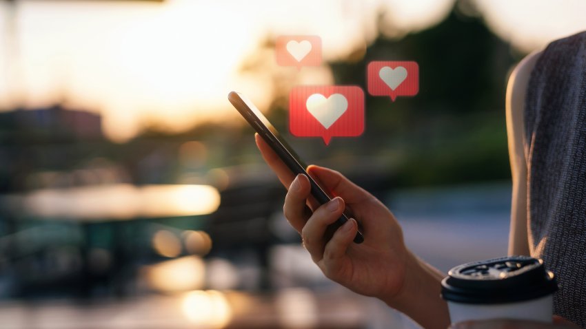 Close up of young woman checking social media network on smartphone on the go in the city. Mobile phone with heart emoji speech bubble message on the screen. People engaging in networking with technology. Social media addiction concept.